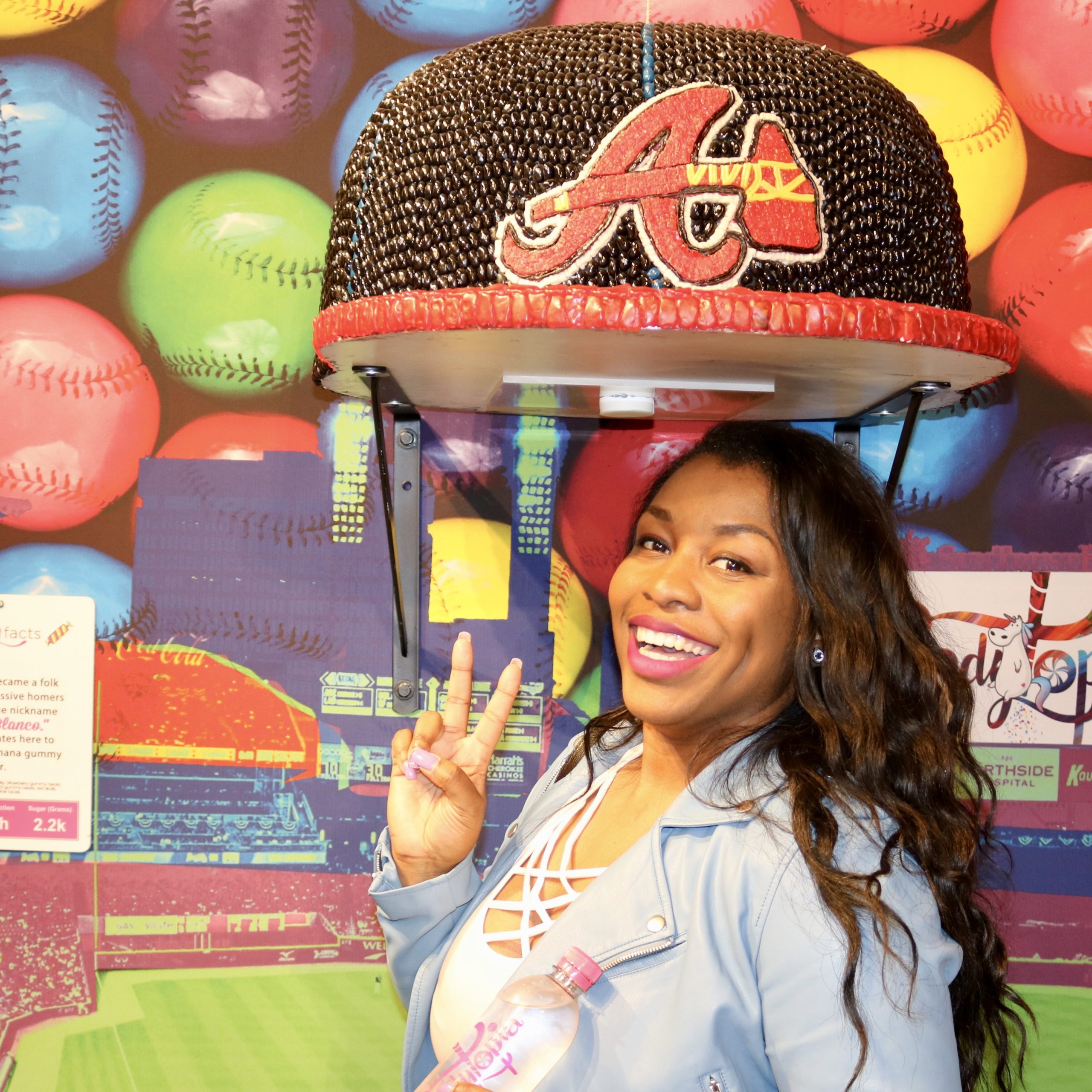 Woman poses under an Atlanta braves baseball hat art piece made out of candy at a museum.