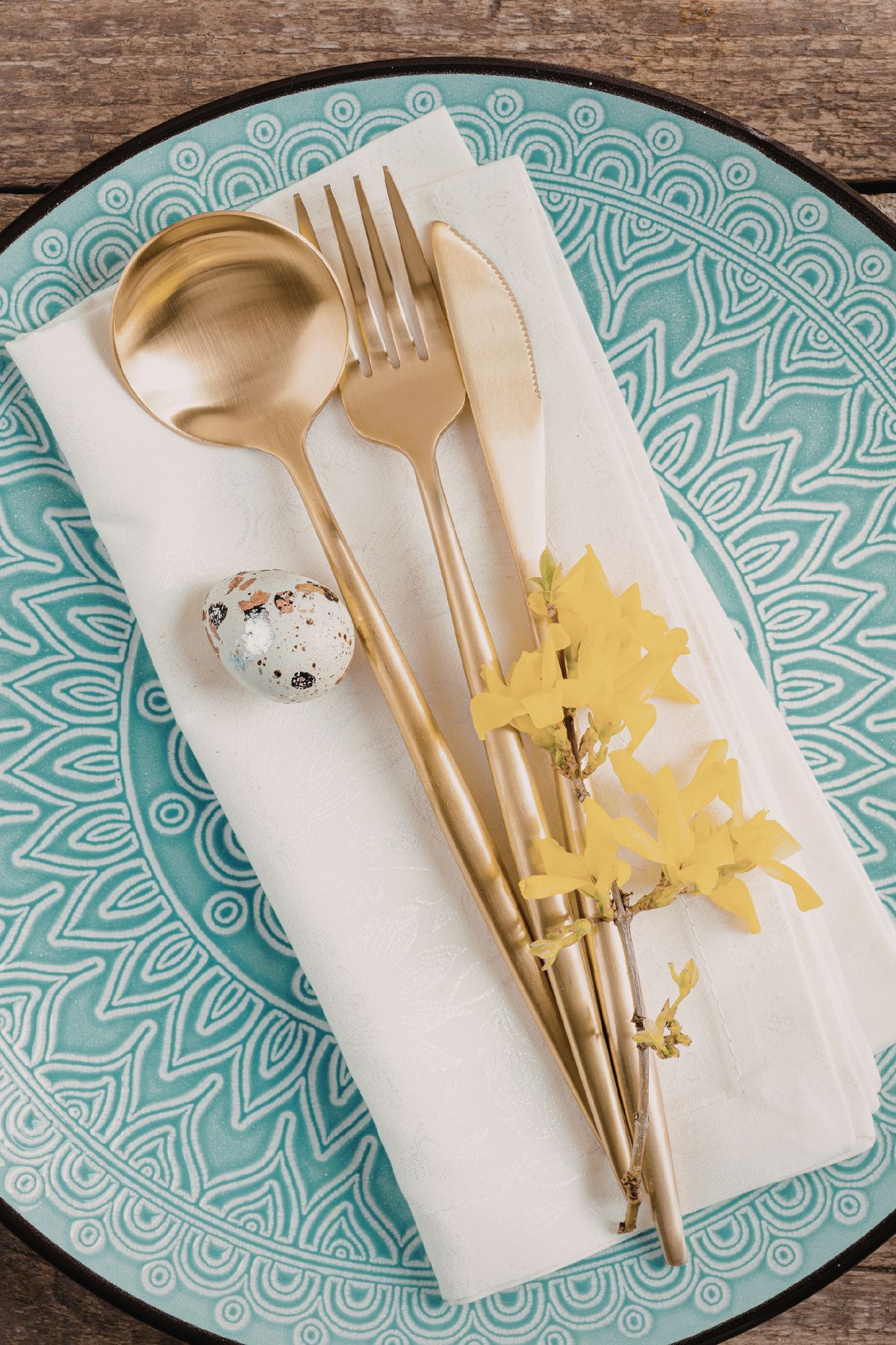 Teal ceramic place with a white napkin, gold utensils, a mini speckled egg, and yellow flowers.