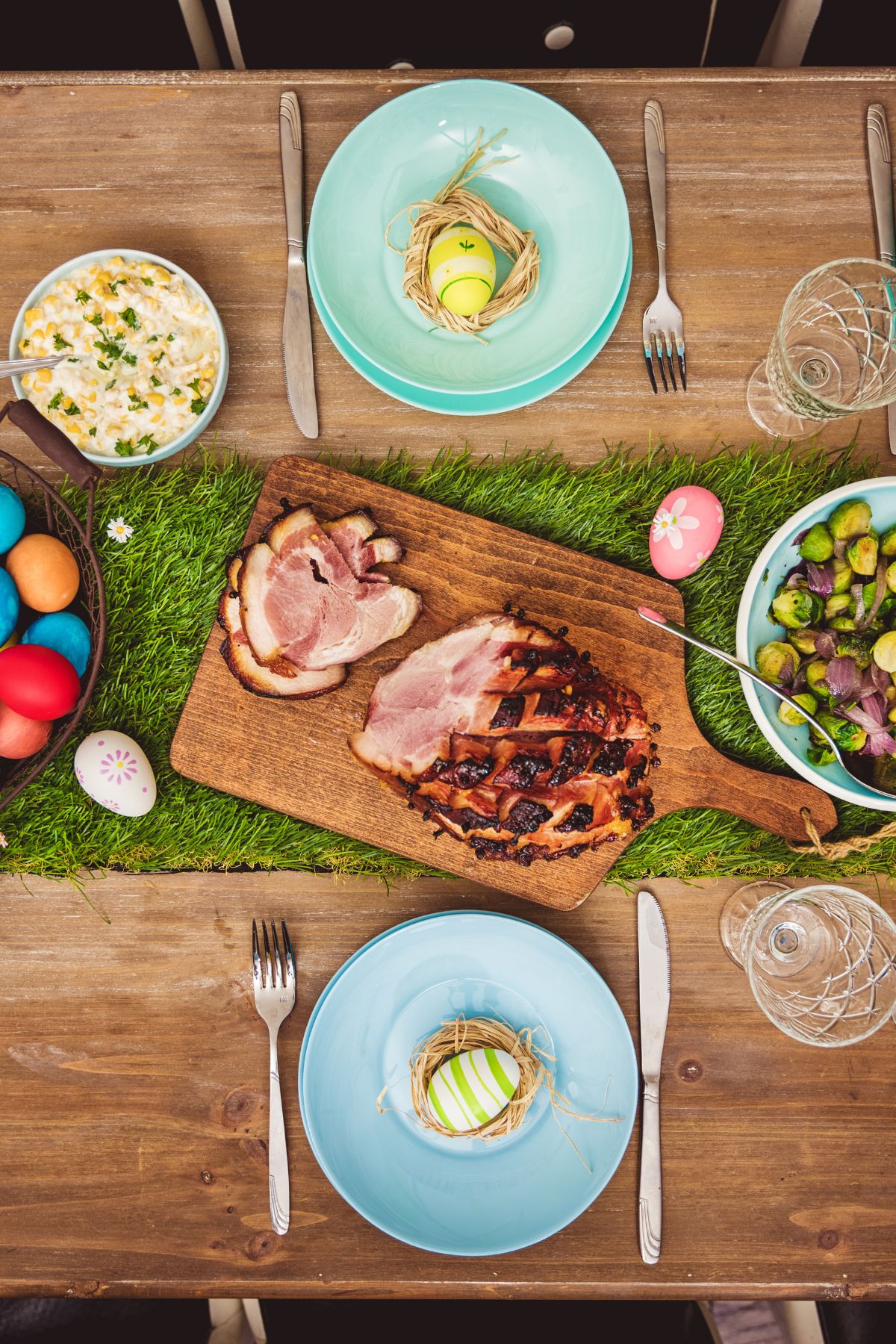 An Easter table with dinner food and teal plates set with colorful eggs wrapped in dried grass.