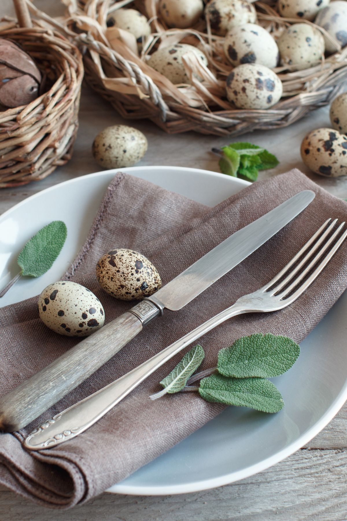 A light blue plate with a gray napkin, utensils, mini speckled eggs, and green leaves for decor.