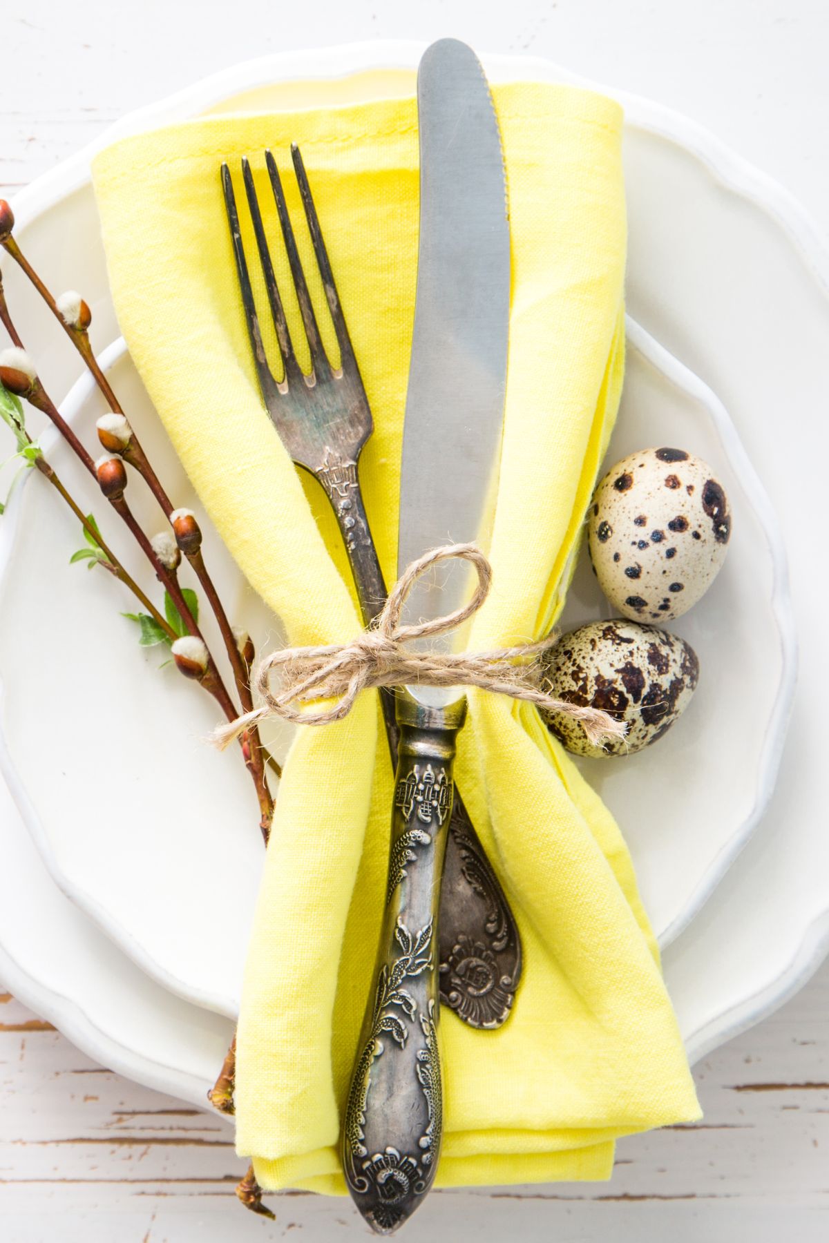 Stacked white plates with a yellow napkin and silverware wrapped in twine with spring foliage and mini speckled eggs.