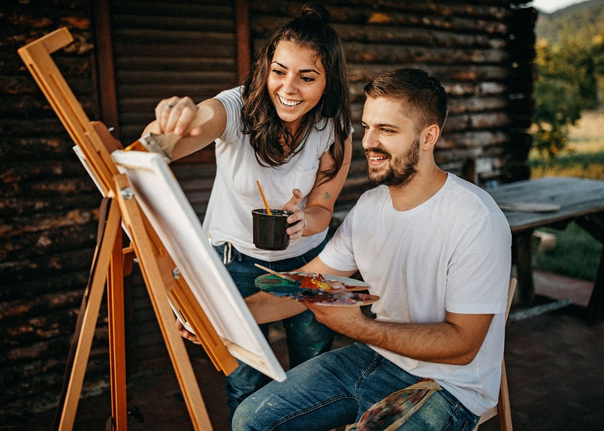 A couple painting on a canvas board outside of a cabin.
